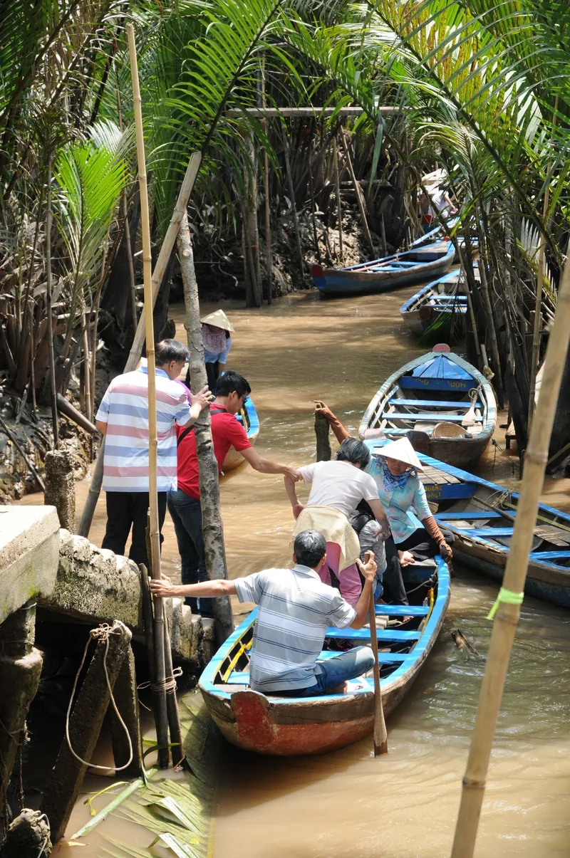 Tourists board small wooden sampans along a narrow canal shaded by palm trees in the Mekong Delta.