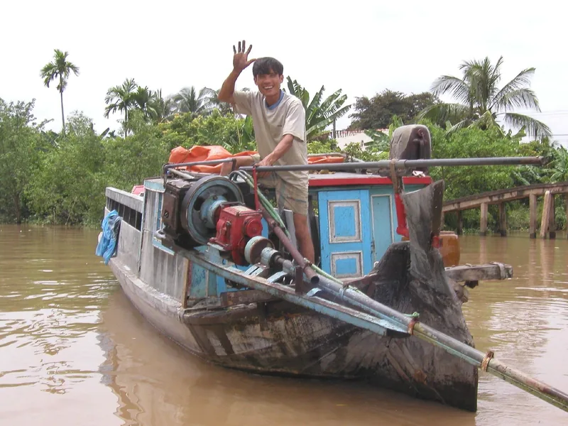 A boatman waves while standing on his wooden engine boat on a muddy river in the Mekong Delta.